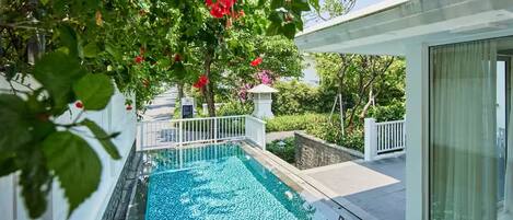 A swimming pool with a white fence, surrounded by lush greenery and a white building with large windows.
