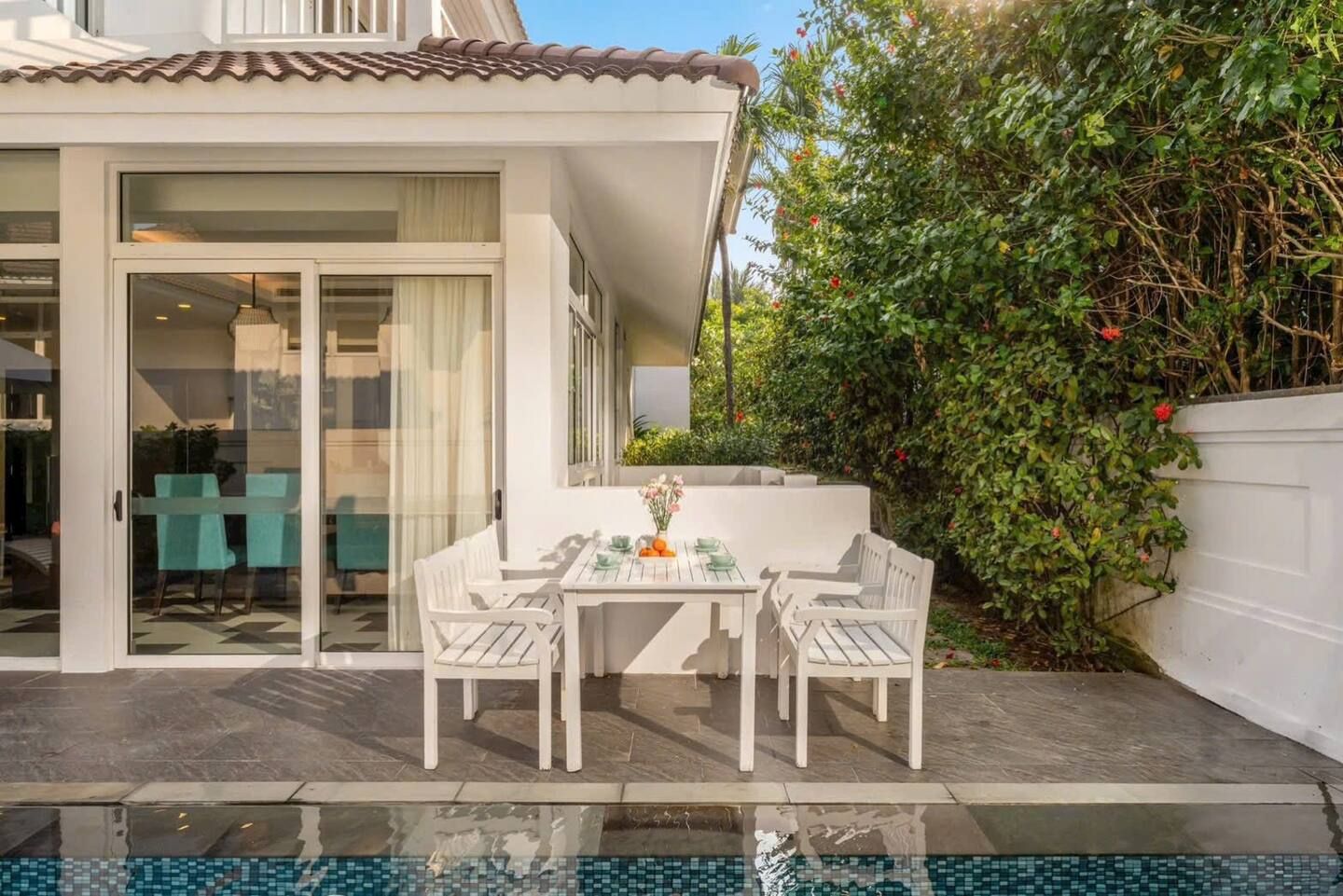 A modern outdoor patio with a glass door leading to a dining area, a poolside table with chairs, and a lush green hedge.