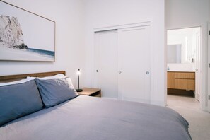 Ein Schlafzimmer mit einem Bett, einem Nachttisch, einer Nachttischlampe, einer Tür und einem durch eine geöffnete Tür einsehbaren Badezimmer.