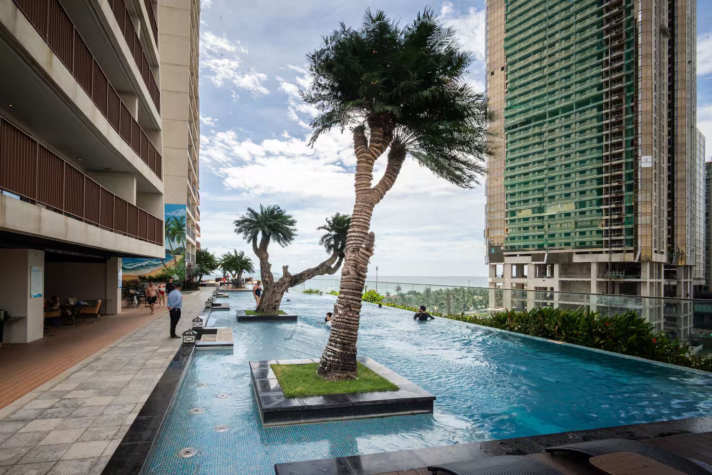 Una piscina con palmeras, un área de restaurante y edificios altos al fondo.