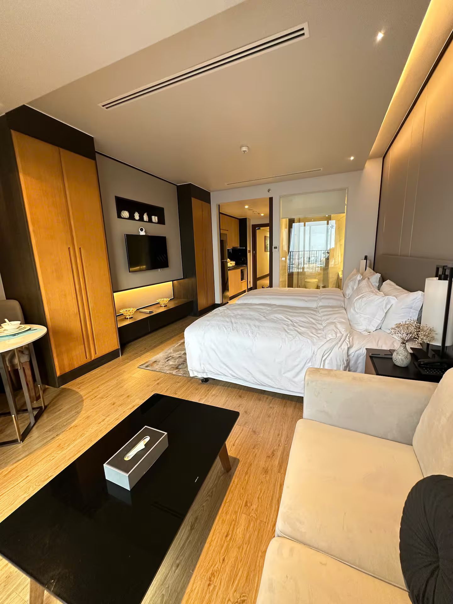 A modern hotel room with a large bed, a sofa, a coffee table, a TV, and a bathroom visible through an open door.