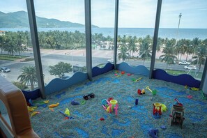 Ein Zimmer mit Blick auf Strand und Palmen, ein Spielbereich mit Spielzeug sowie ein Fenster mit Blick auf eine Straße und Fahrzeuge.