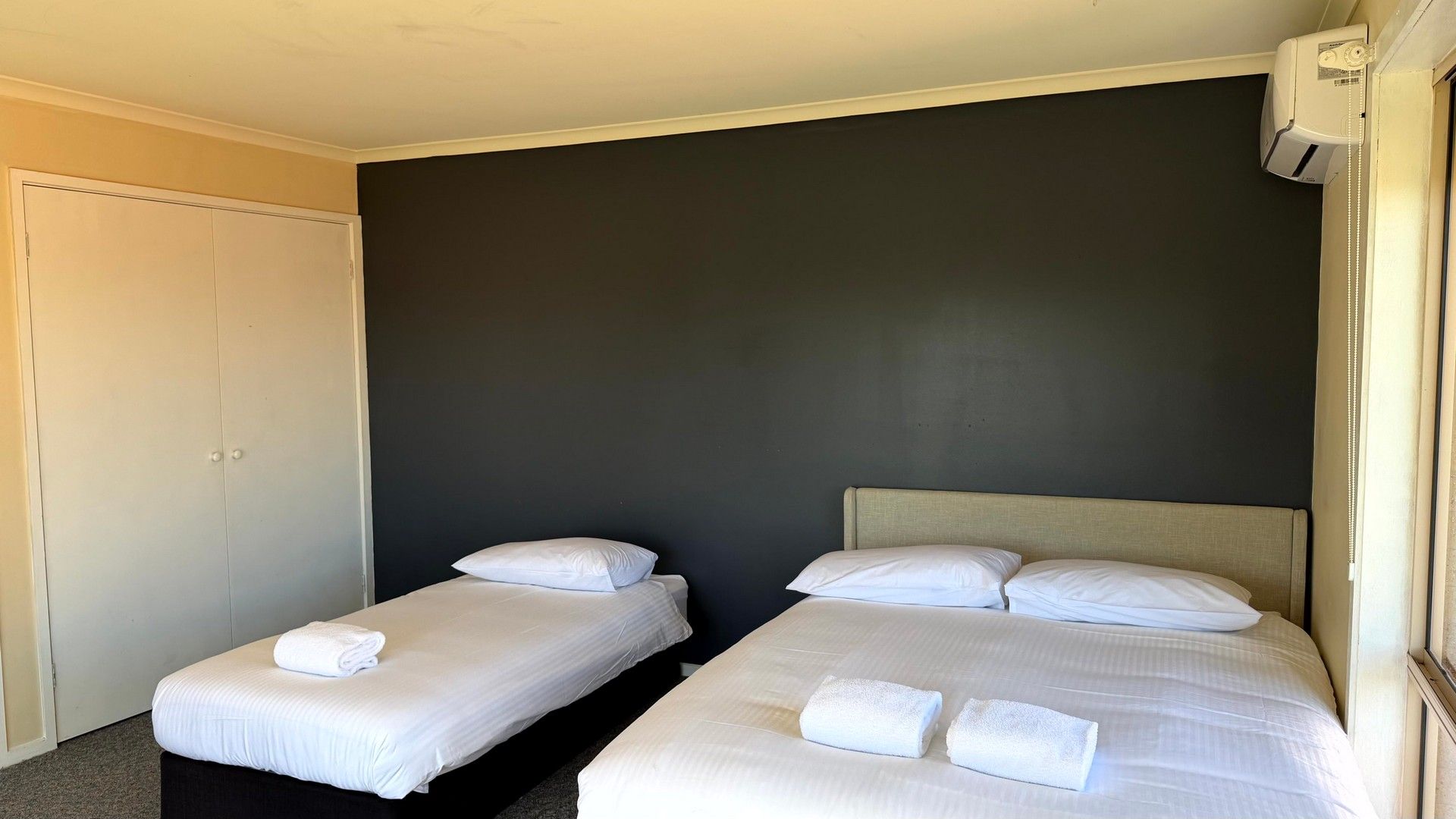 A hotel room with two single beds, a wardrobe, and an air conditioning unit.