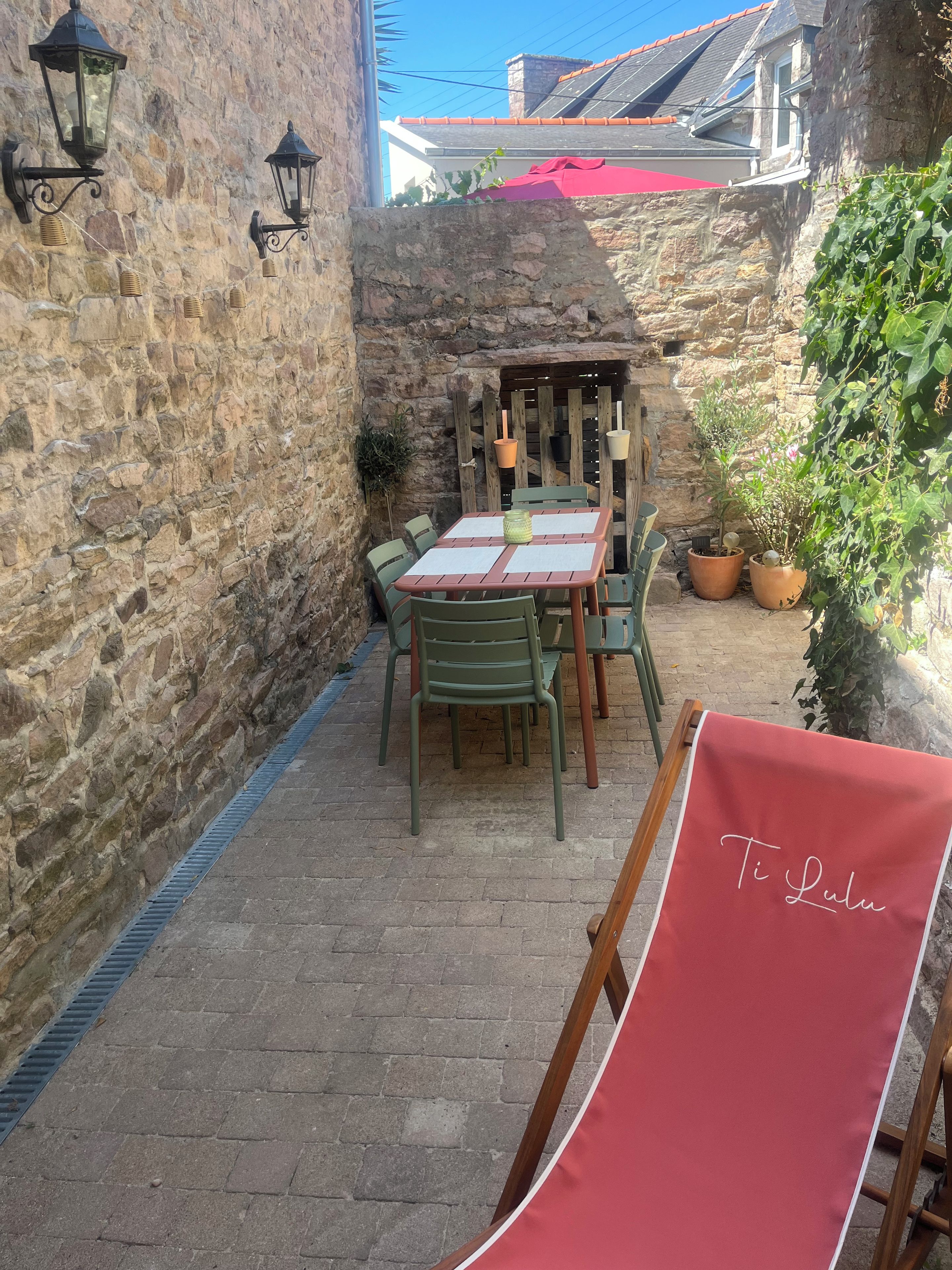 A stone wall with a wooden table and chairs, a red cloth, and potted plants.