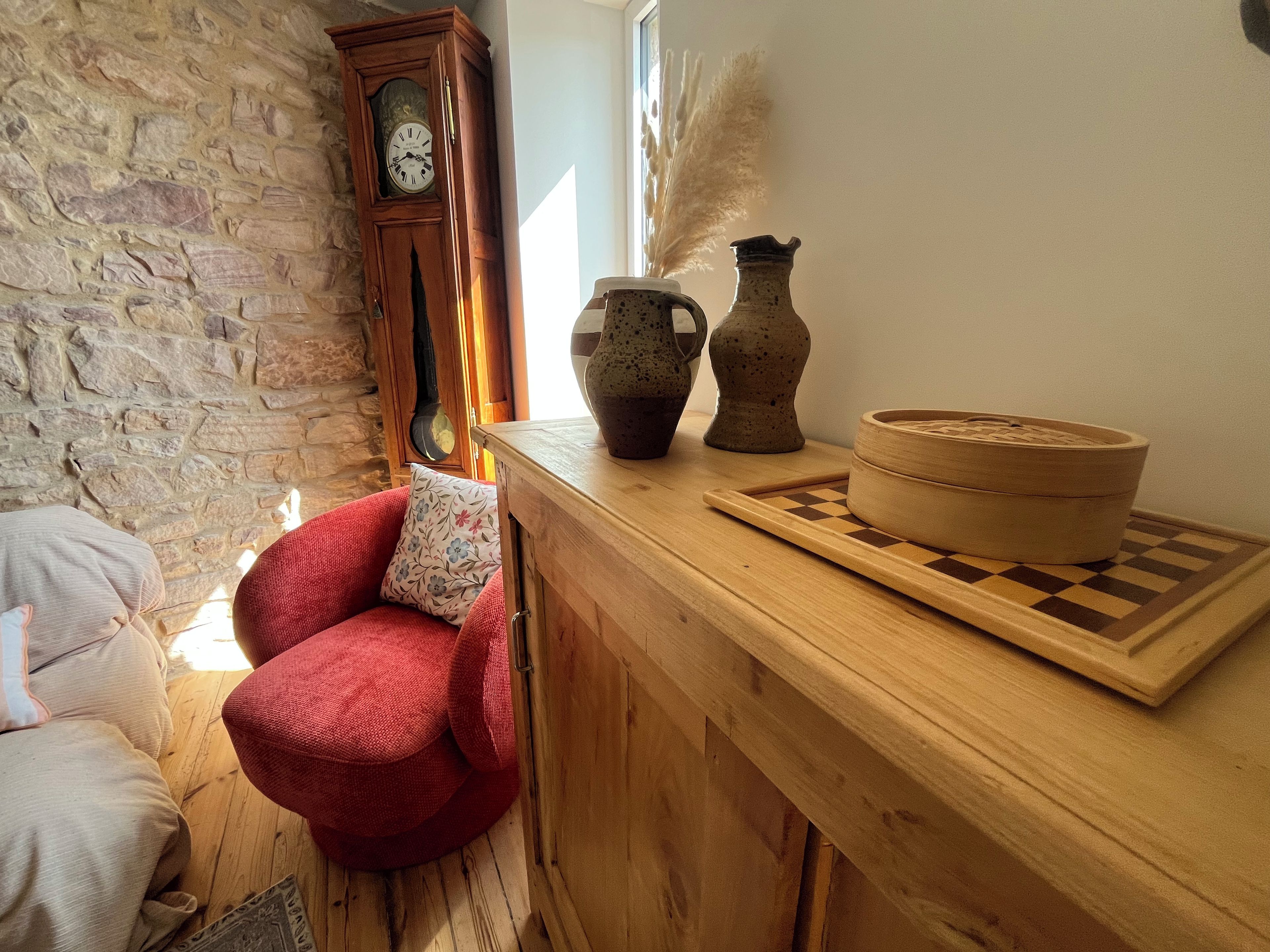 A cozy room with a stone wall, a wooden clock, a red chair, and a wooden table with a chessboard and a woven basket.
