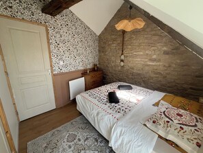 A bedroom with a sloped ceiling, a bed with patterned bedding, a wooden dresser, a radiator, and a decorative wall. - The Little Pink Sandstone House “Ti Lulu”—A charming fisherman’s cottage with a view of the sea (Erquy)