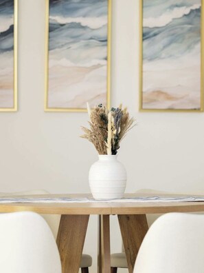 Une table à manger surmontée d’un vase blanc contenant des plantes séchées, le tout placé devant un mur orné de trois tableaux abstraits encadrés.