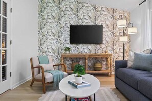A living room with a flat-screen TV mounted on the wall, a round coffee table, a sofa, a chair, and a console table with a plant. - Players Club Getaway I Sawgrass (Palm Valley)