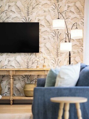 Sala de estar com televisão de tela plana na parede, papel de parede com padrão de folhas, mesa de centro de madeira e sofá azul com almofadas.