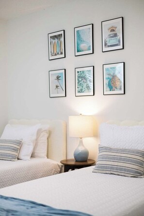 Quarto com duas camas, mesa de cabeceira com abajur e seis quadros na parede.