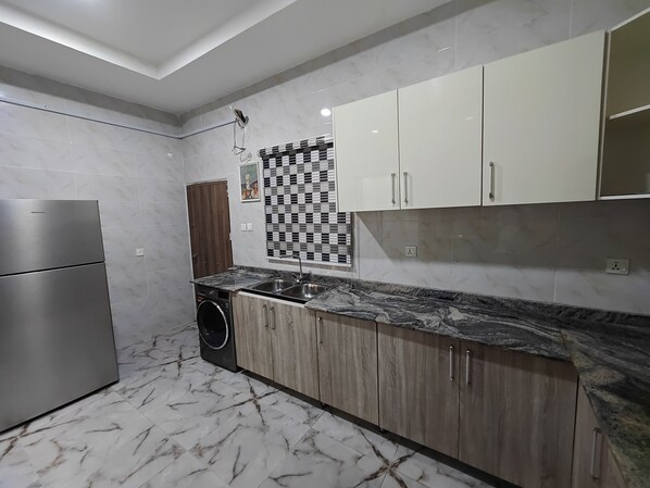 A modern kitchen with a stainless steel refrigerator, wooden cabinets, a built-in washing machine, and a marble-patterned floor.