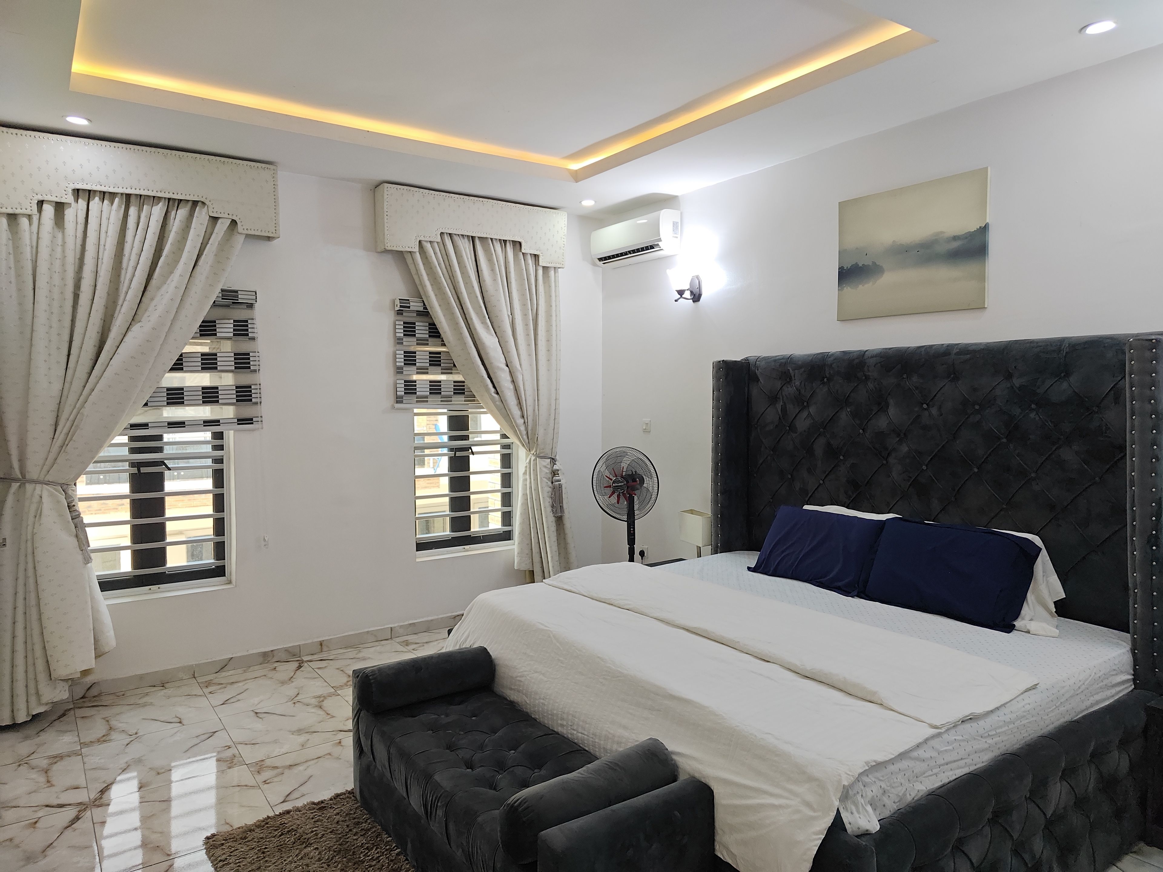Une chambre à coucher avec un grand lit, une table de chevet, un ventilateur sur pied et une fenêtre avec des rideaux.