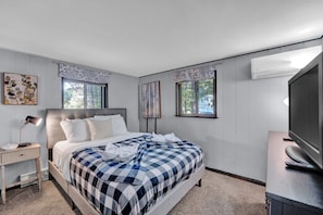 A bedroom with a bed, bedside table, lamp, artwork, windows, and an air conditioning unit. - Hot Tub Cabin Near Skiing & Pocono Raceway. Sleeps 5, Firepit, AC (Long Pond)