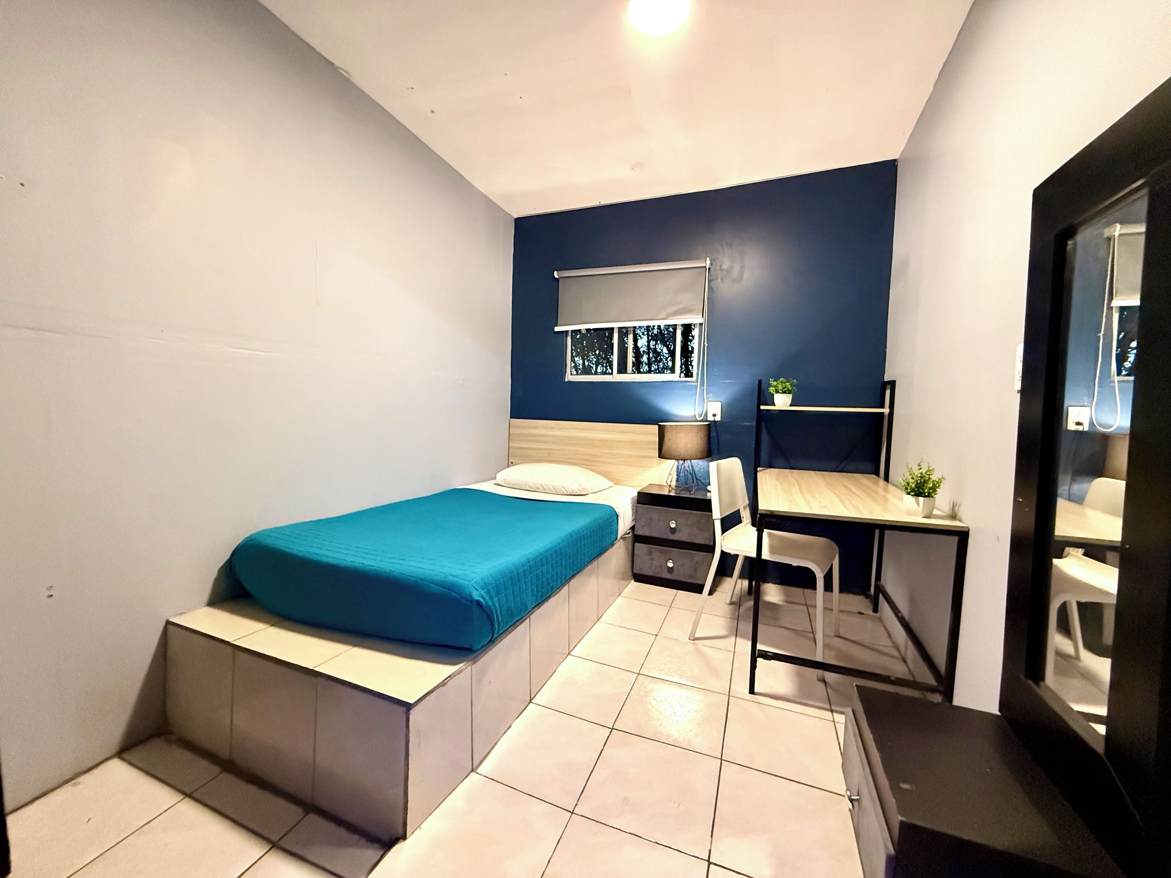 Un dormitorio pequeño con cama, mesita de noche, escritorio, silla y un ventanal.