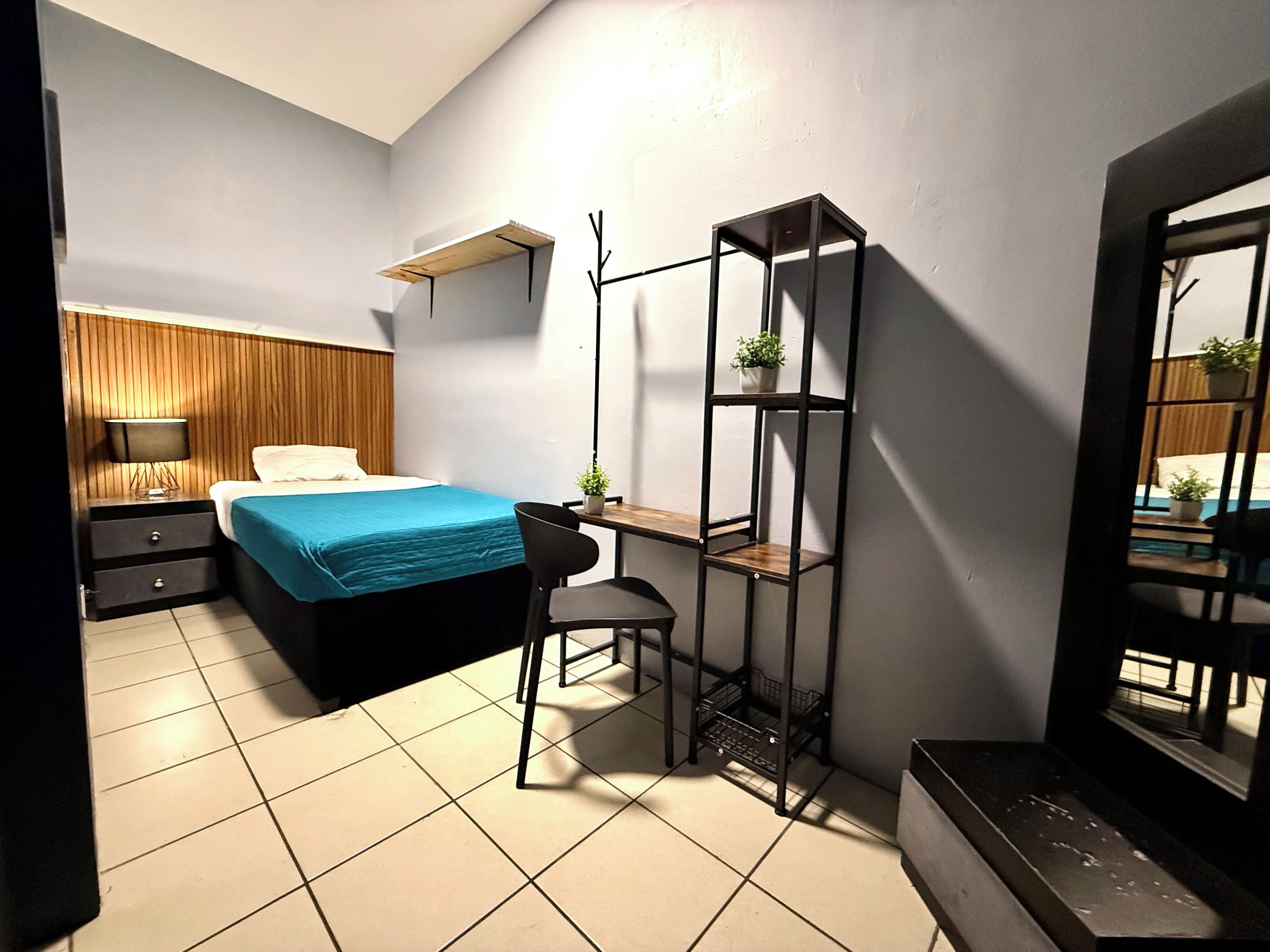 A bedroom with a bed, a desk, a chair, a shelf with plants, and a window.