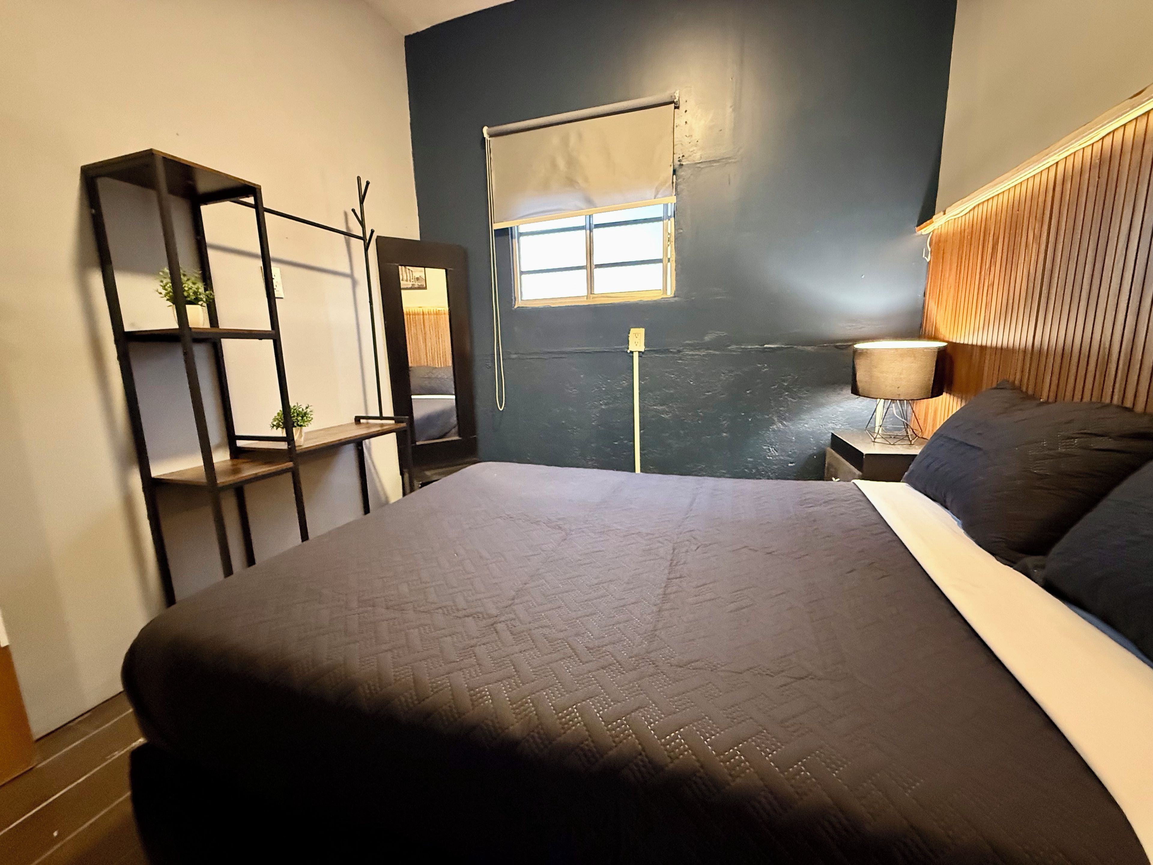 Een slaapkamer met een groot bed, een nachtkastje met een lamp, een raam met een blind, een wandplank met planten, en een deur met een handdoekhouder.