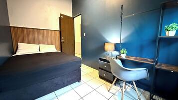 A bedroom with a bed, a desk with a chair, a lamp, and a shelf with a plant.