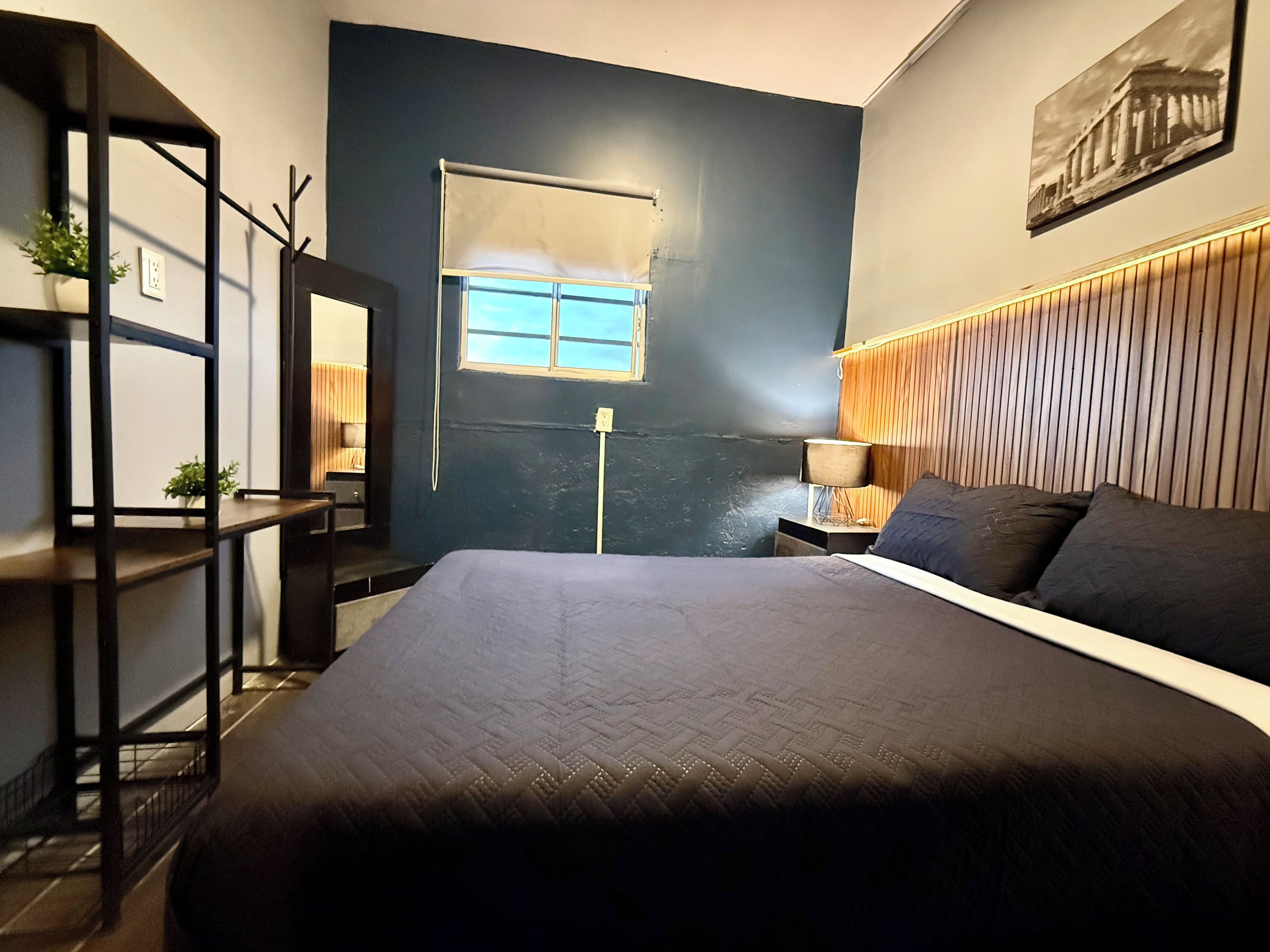 A bedroom with a bed, a nightstand, a lamp, a window, and a wall-mounted shelf with plants.