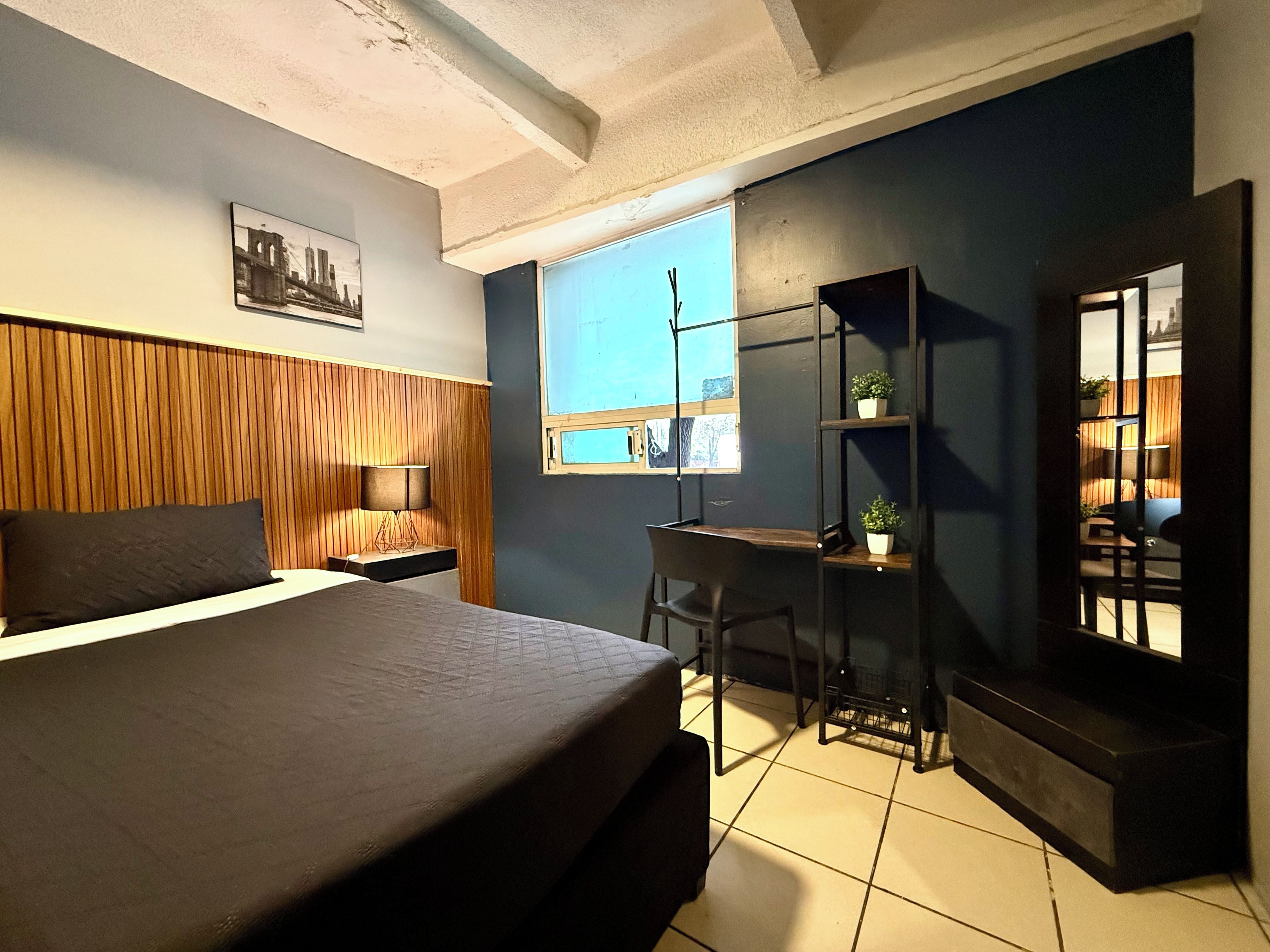 A bedroom with a bed, a desk, a chair, a window, and a shelf with plants.