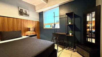 A bedroom with a bed, a desk, a chair, a window, and a shelf with plants.