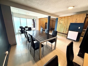 Uma área de jantar moderna com uma mesa comprida e cadeiras, um painel de madeira decorativo e uma pequena cozinha compacta.