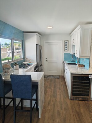 A modern kitchen with white cabinets, a stainless steel refrigerator, a dining area with blue chairs, and a view of the outdoors through a window. - Coastal 1 Bedroom Unit with 40 foot boat slip (Tavernier)