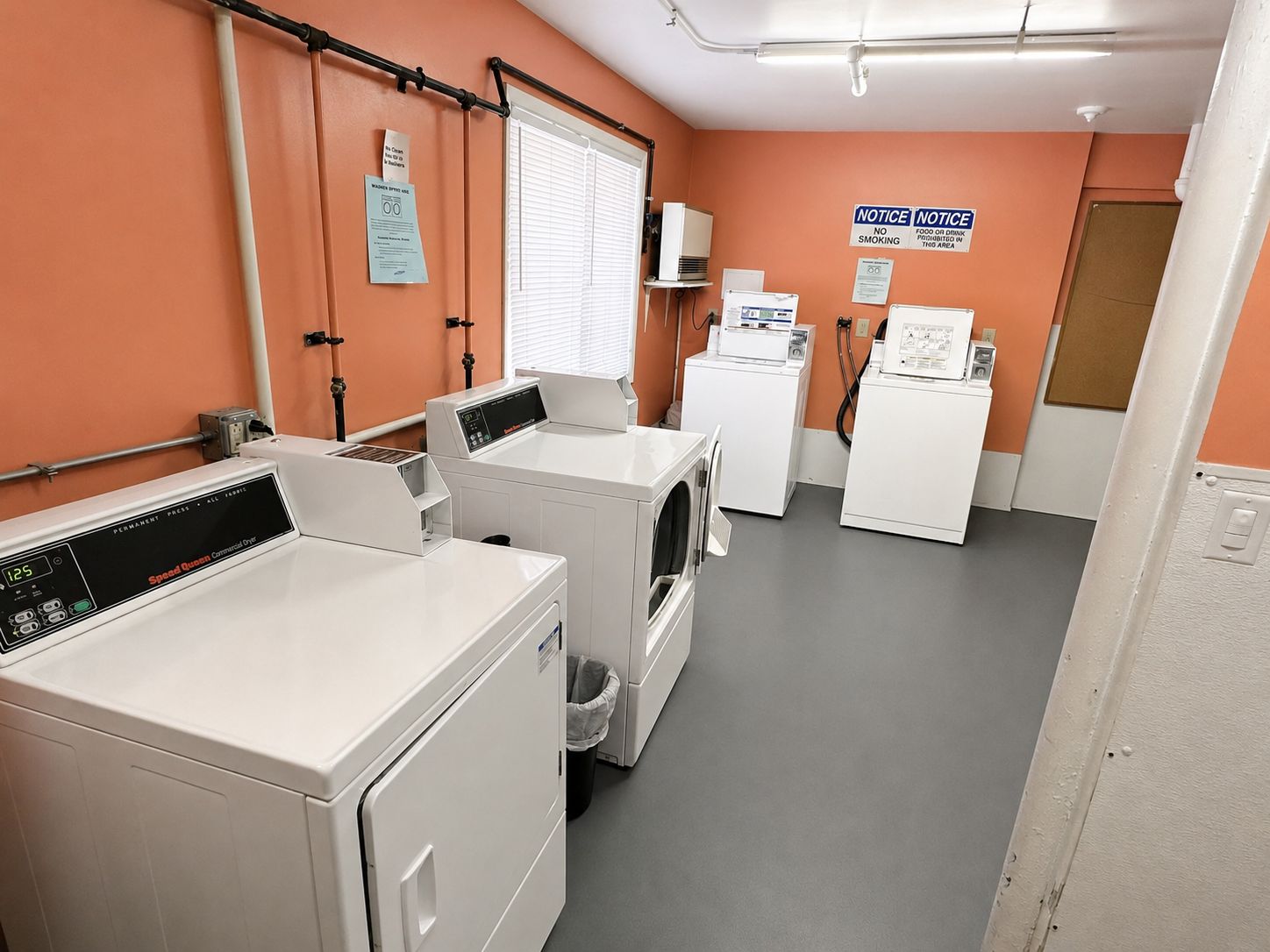 Une buanderie équipée de plusieurs machines à laver et de plusieurs sèche-linge, un panneau d’affichage et une fenêtre avec des stores.