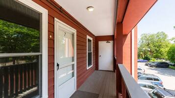 A covered porch with a white door, a window with a screen, and a view of parked cars and trees.