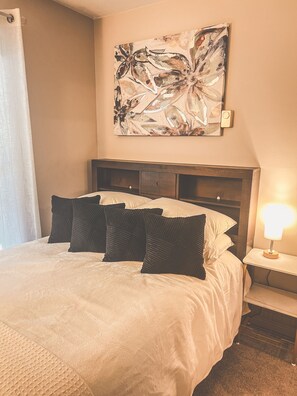 A hotel room with a bed, a nightstand with a lamp, and a wall-mounted abstract painting. - Downtown 2BR–Ideal Mid-Term Stay Near UVM & UVMMC (Burlington)