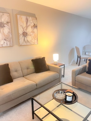 Un salon moderne avec un canapé beige, une table basse en verre et une table d’appoint avec une lampe. Deux tableaux abstraits encadrés ornent le mur.