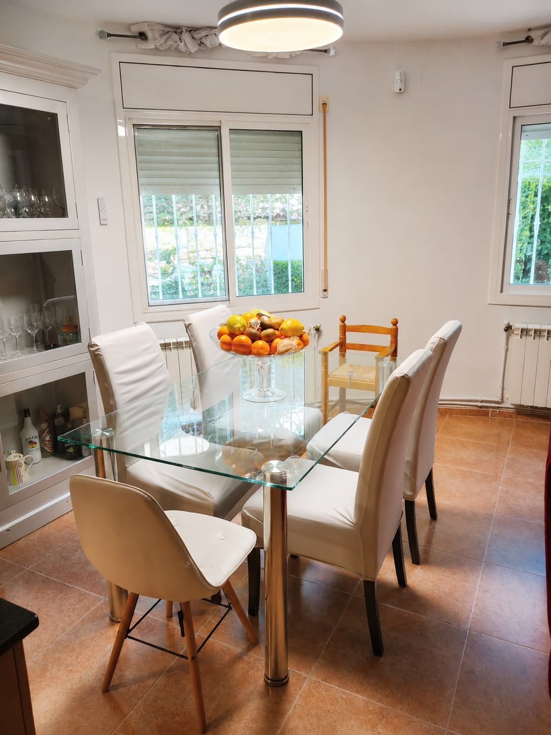 A dining area with a glass table surrounded by chairs, a bowl of fruit, and a view of the outdoors through the windows.