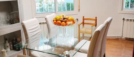 A dining area with a glass table surrounded by chairs, a bowl of fruit, and a view of the outdoors through the windows.