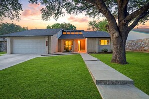 A residential house with a garage, a well-maintained lawn, and a large tree in the front yard. - Resort Getaway: Pool, Hot Tub & Putting Green (San Antonio)