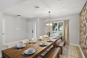 A dining area with a long wooden table set for six, a hanging light fixture, and a view through a window with curtains. - Resort Getaway: Pool, Hot Tub & Putting Green (San Antonio)