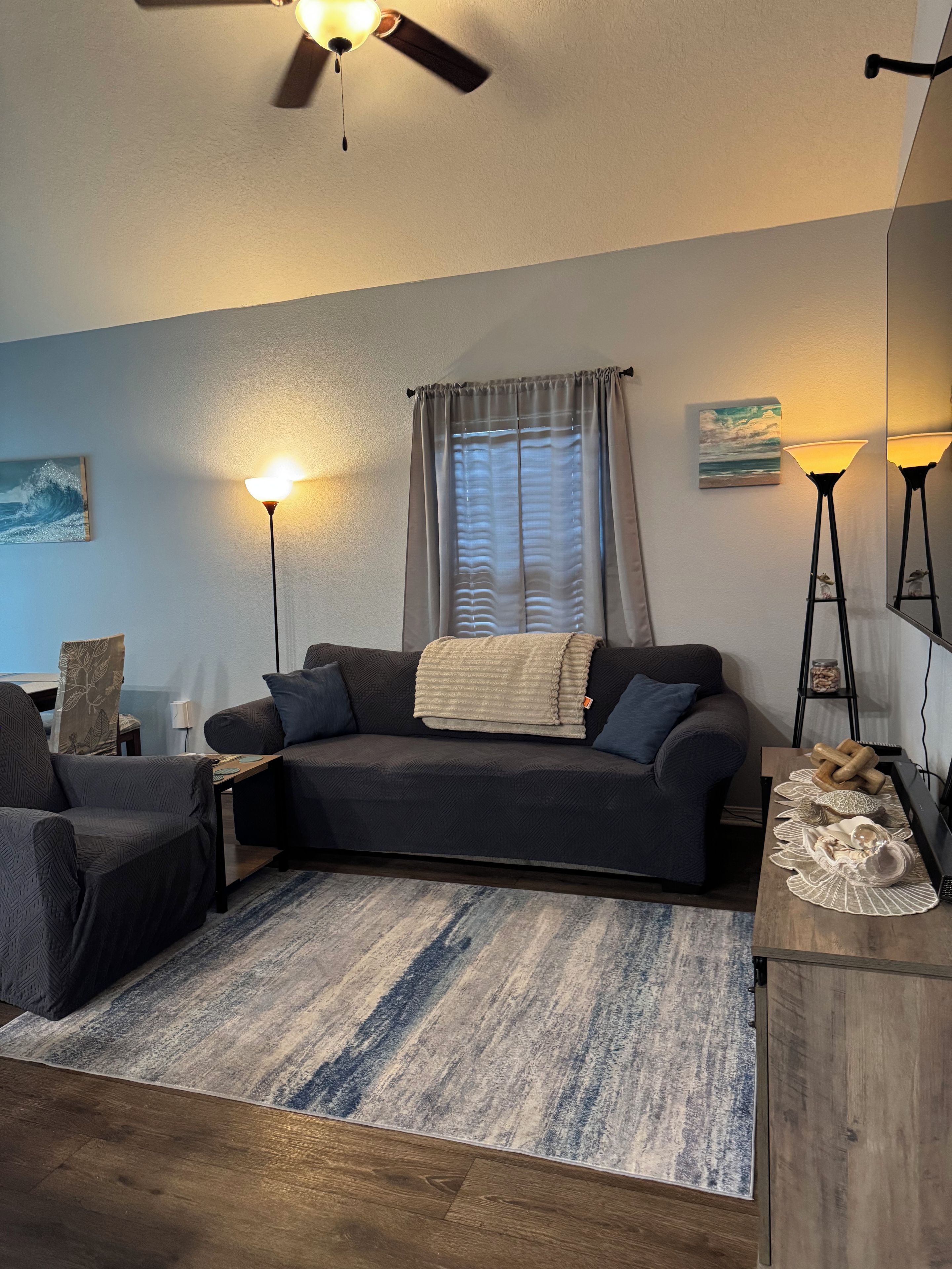 A living room with a dark-colored sofa, a coffee table, a floor lamp, a ceiling fan, and a window with curtains.