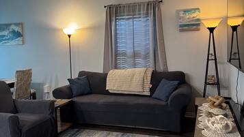 A living room with a dark-colored sofa, a coffee table, a floor lamp, a ceiling fan, and a window with curtains.