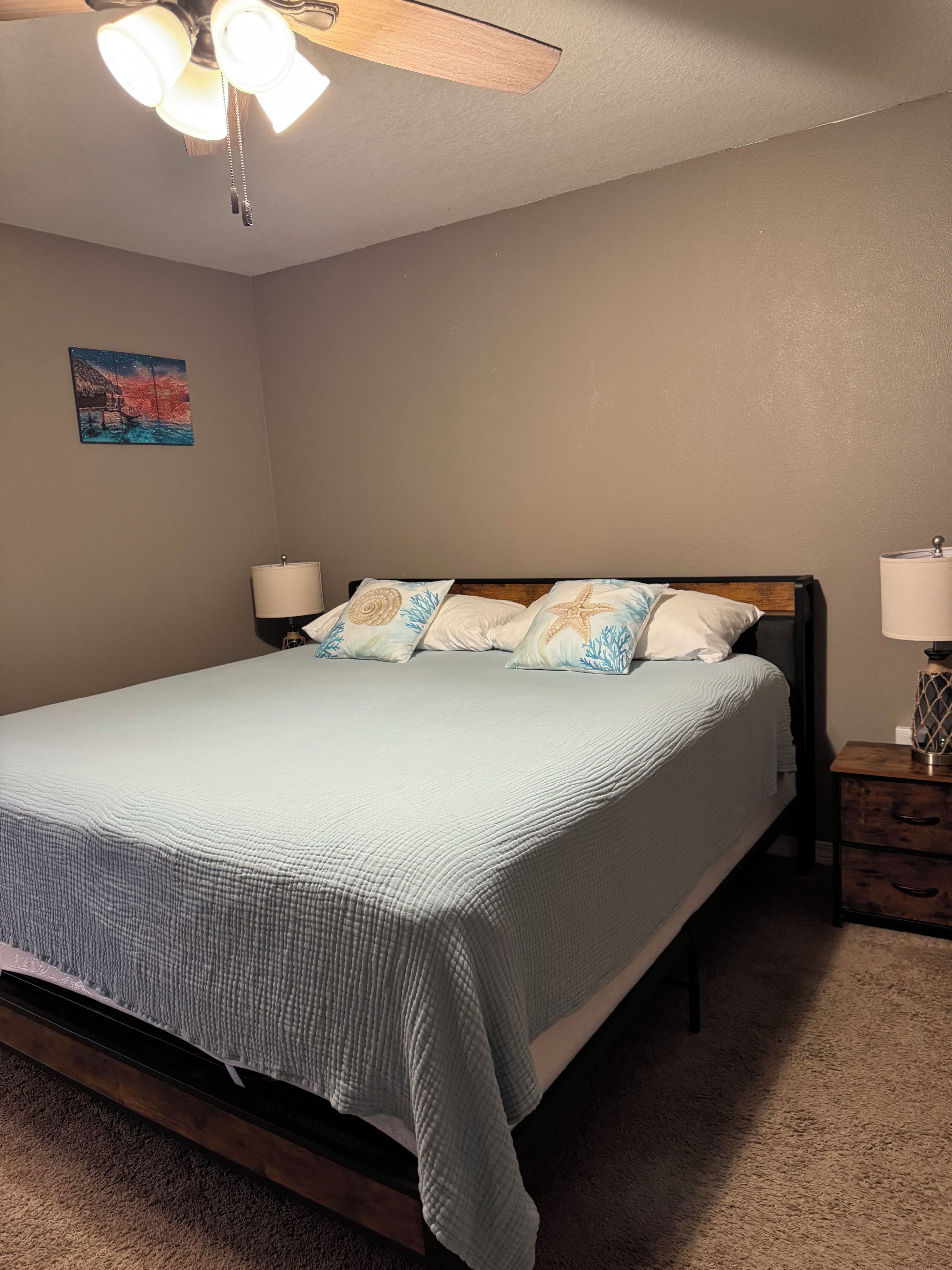 A bedroom with a bed, a nightstand, a lamp, a ceiling fan, and a wall painting.
