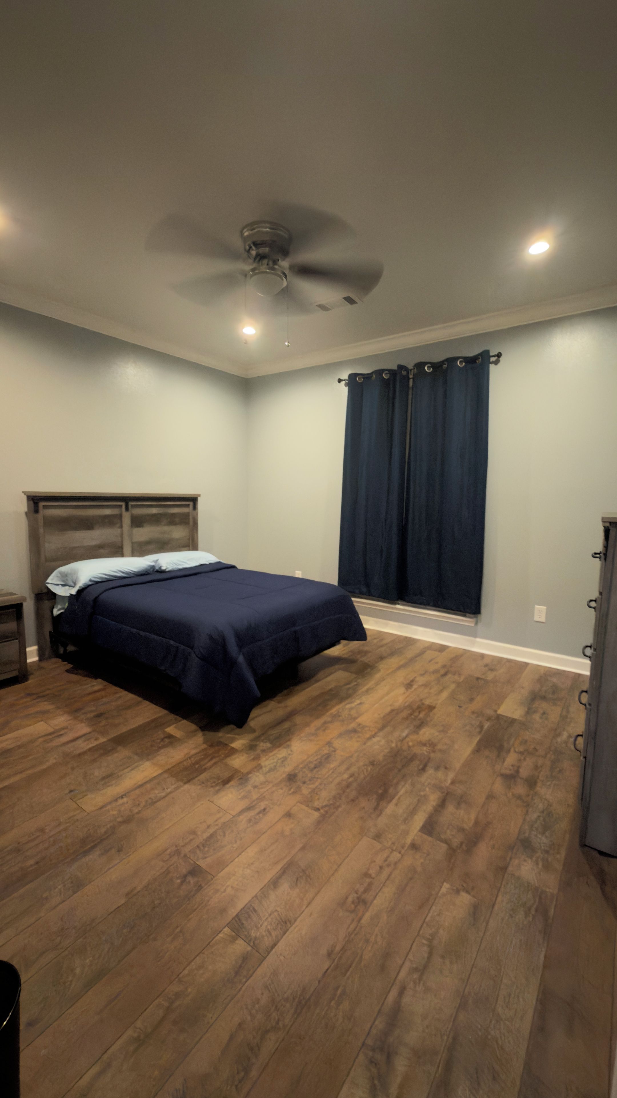 Une chambre à coucher avec un lit, un ventilateur de plafond, une fenêtre avec des rideaux et un plancher en bois.