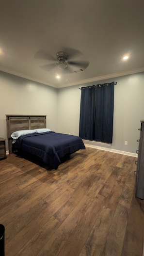 Quarto com cama, ventilador de teto, janela com cortinas e piso de madeira.