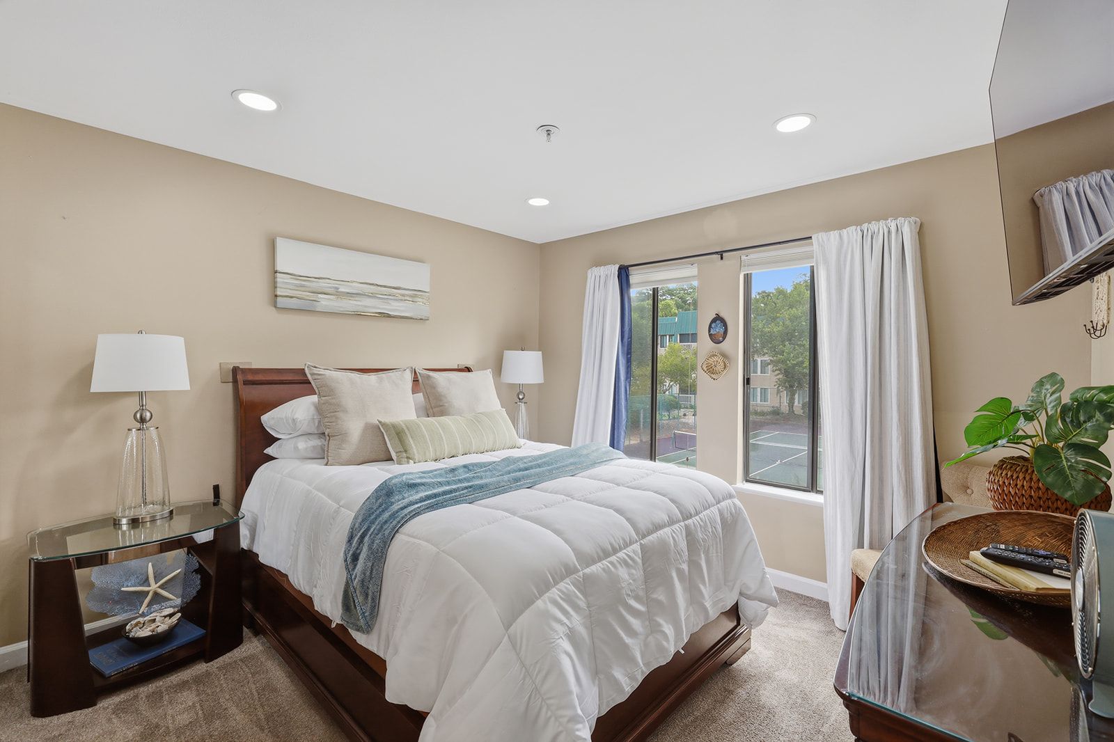 A hotel room with a bed, bedside lamps, a window with curtains, a view of the outdoors, and a plant on a side table.