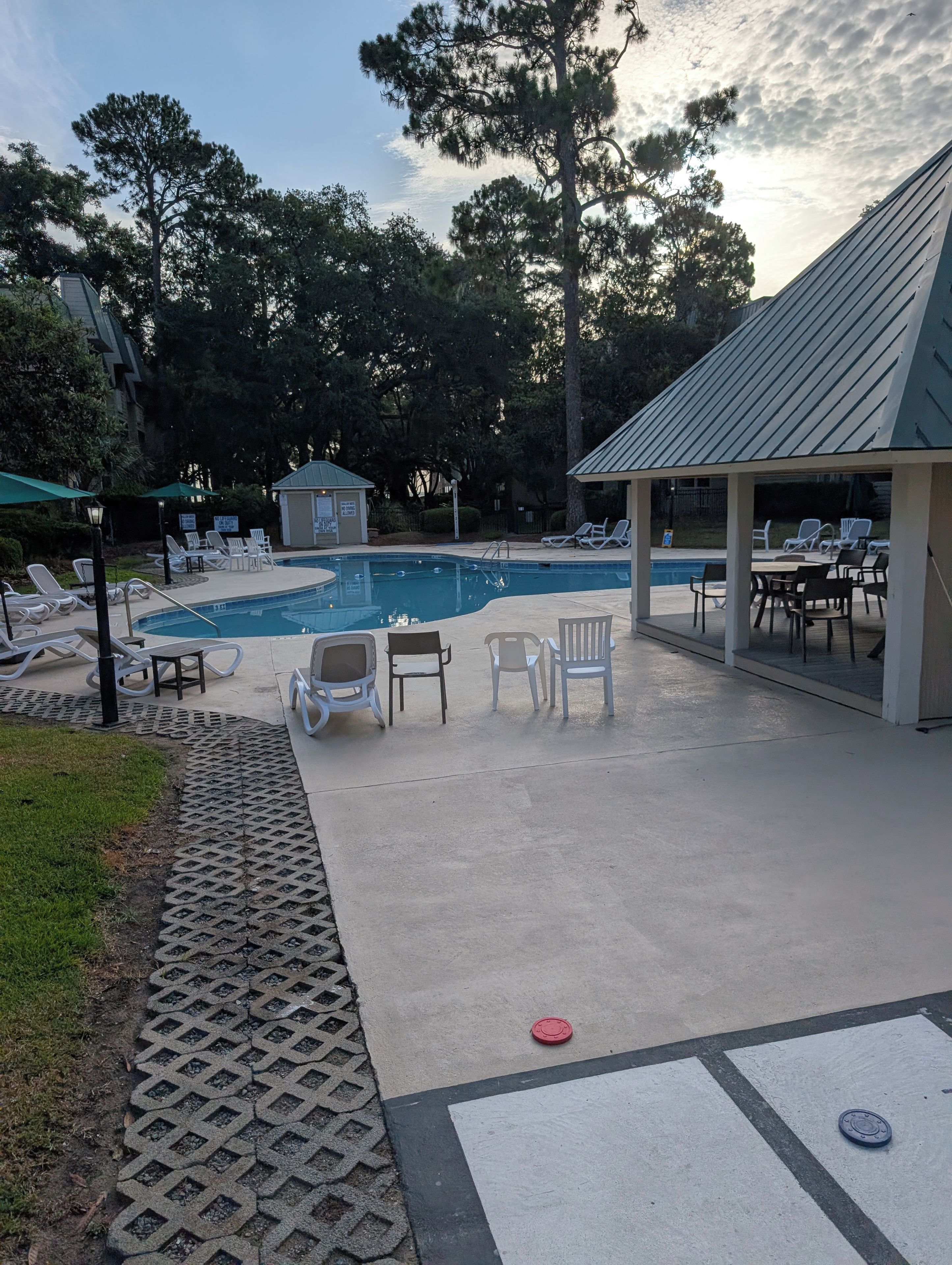Un área de piscina con sillas de descanso, un espacio de estar cubierto y un pequeño edificio.