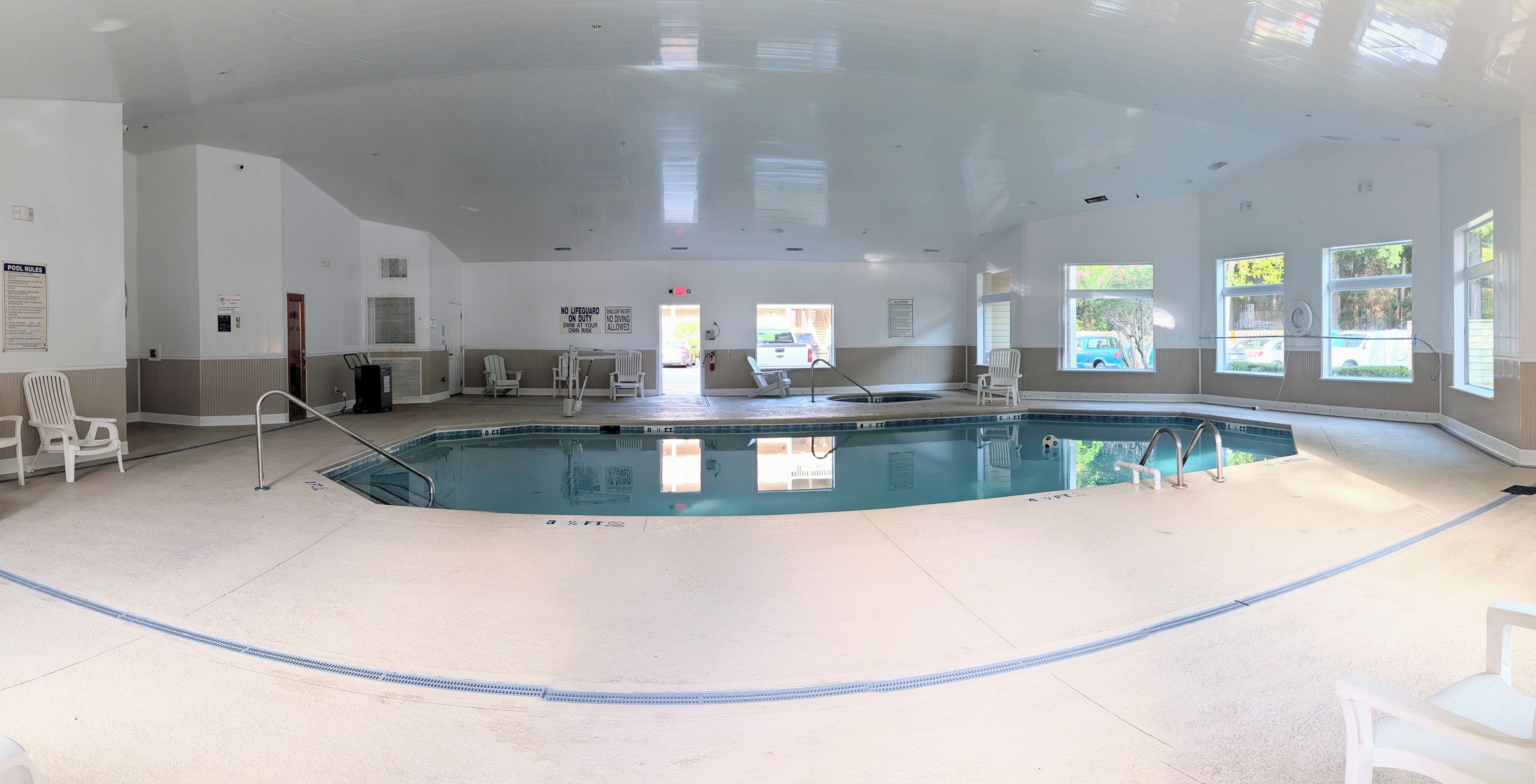 An indoor swimming pool with clear pool rules posted on the wall, a lifeguard chair, and a poolside area with chairs and a sunshade.