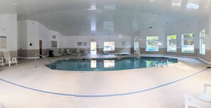 En indendørs swimmingpool med tydelige bassinregler opslået på væggen, en redningsbåd og et område ved poolen med stole og parasol.