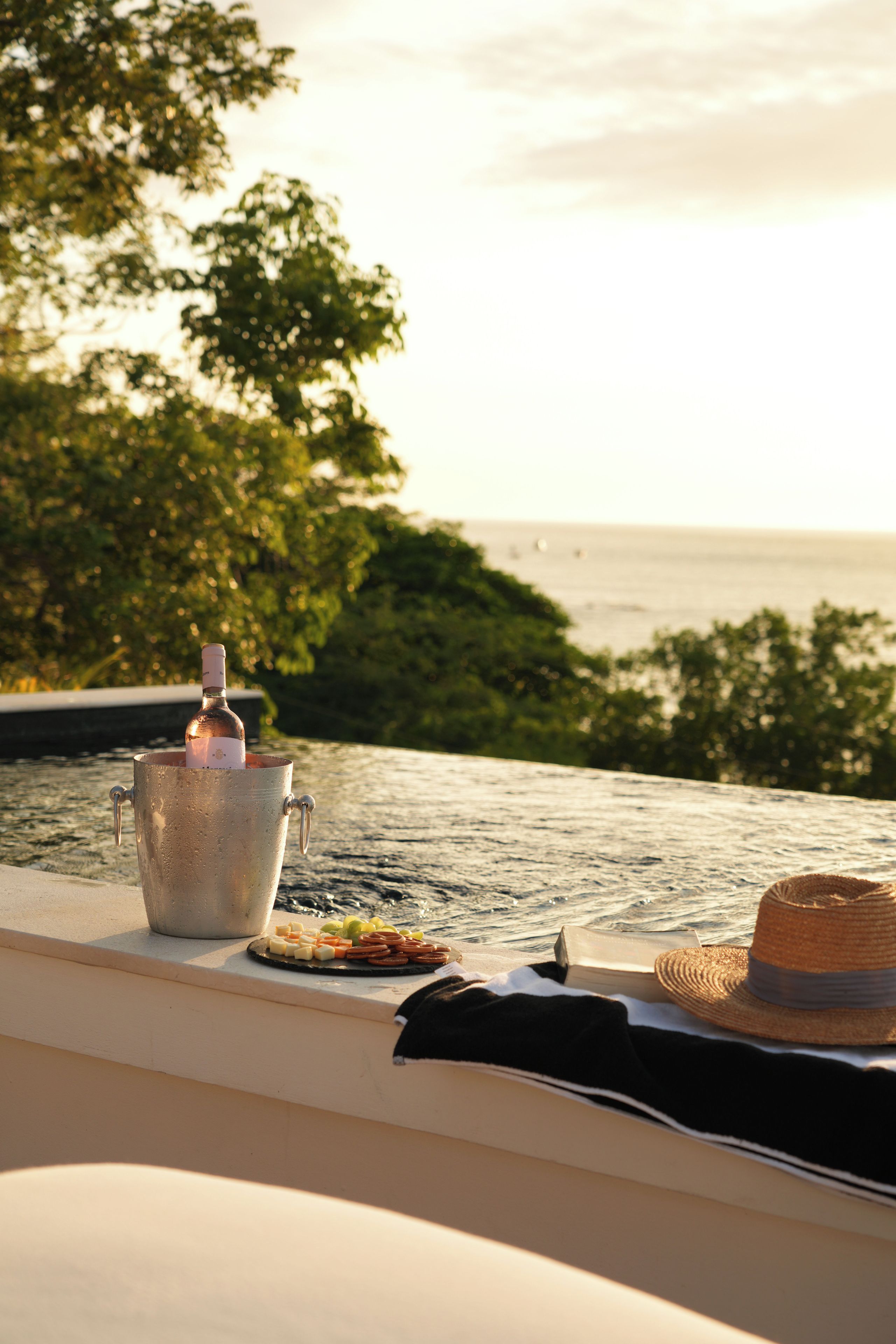 Un balde de metal con una botella, un plato de aperitivos y un sombrero de paja en un balcón con vistas al agua y la vegetación.