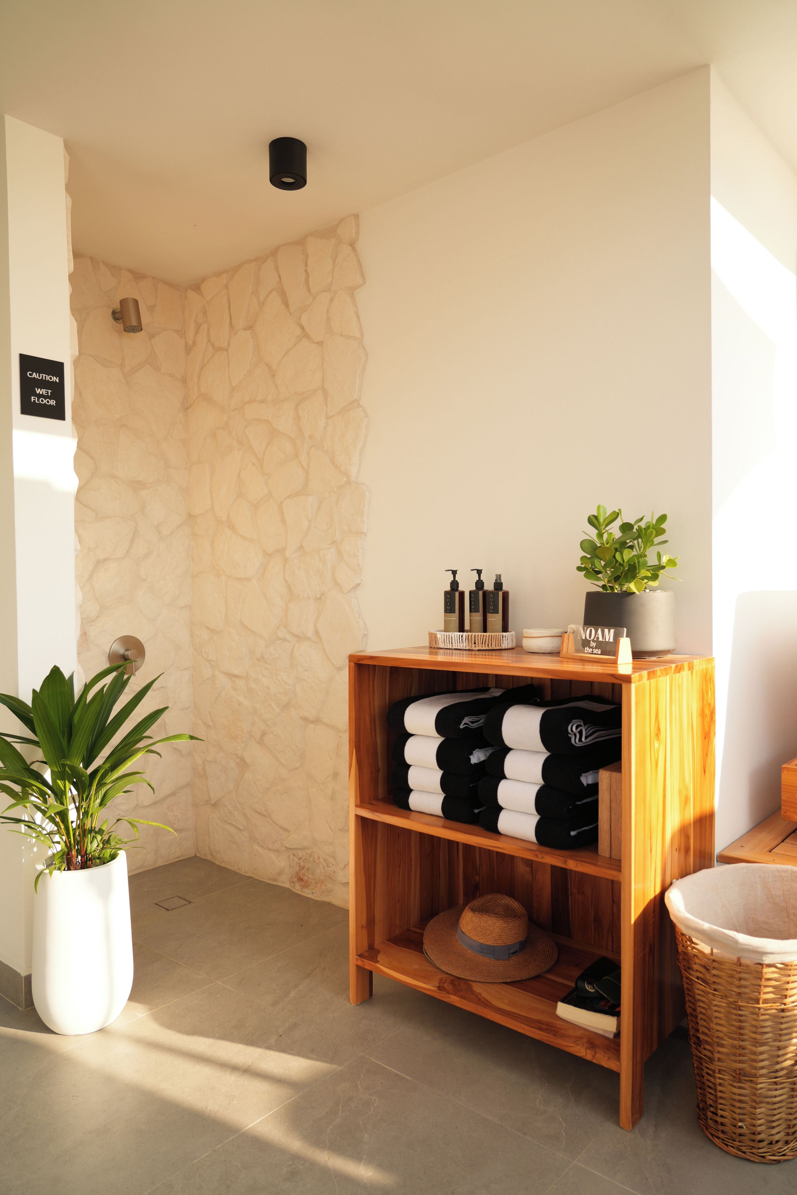Una habitación con pared de piedra, estante de madera con toallas y artículos de aseo, planta en maceta y señal de precaución en el suelo.