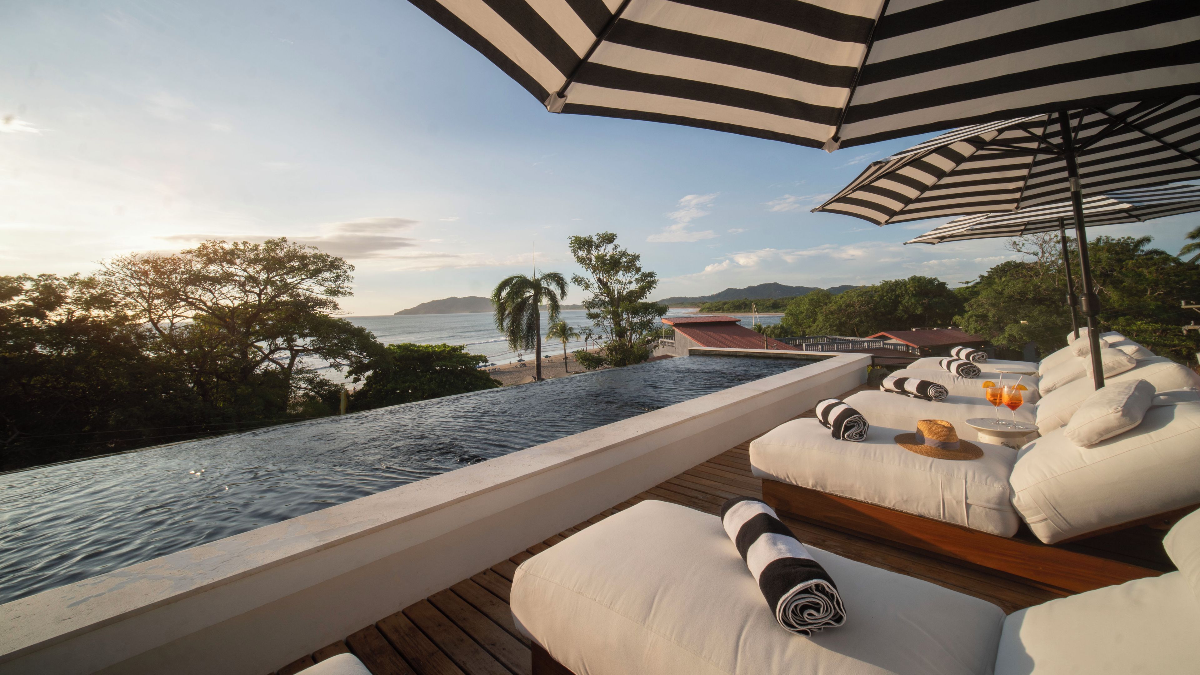 Zona de piscina con sillones de descanso, una sombrilla a rayas y vistas al mar y las colinas.