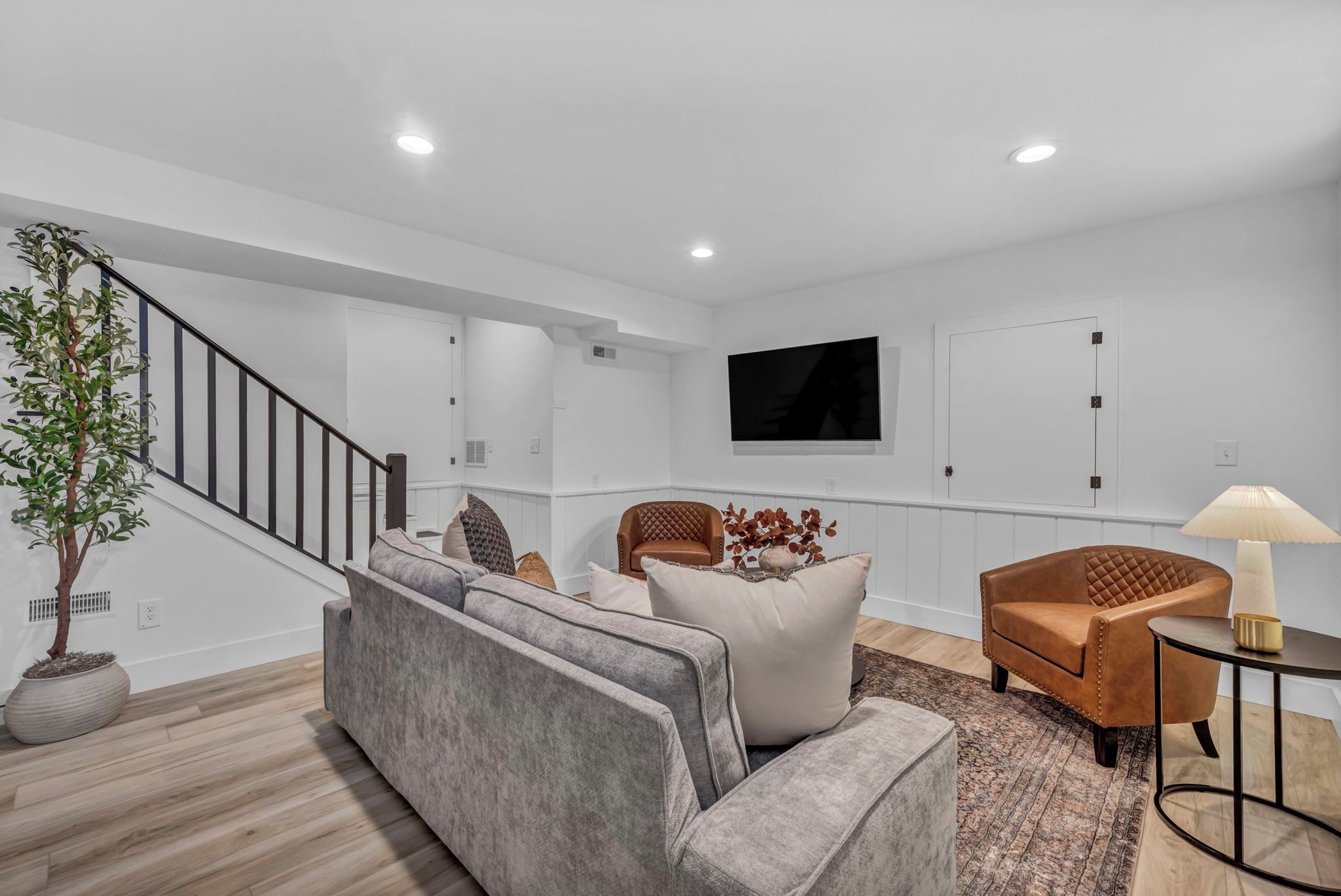Un salon moderne avec un canapé gris, un fauteuil marron, un téléviseur fixé au mur et un escalier.