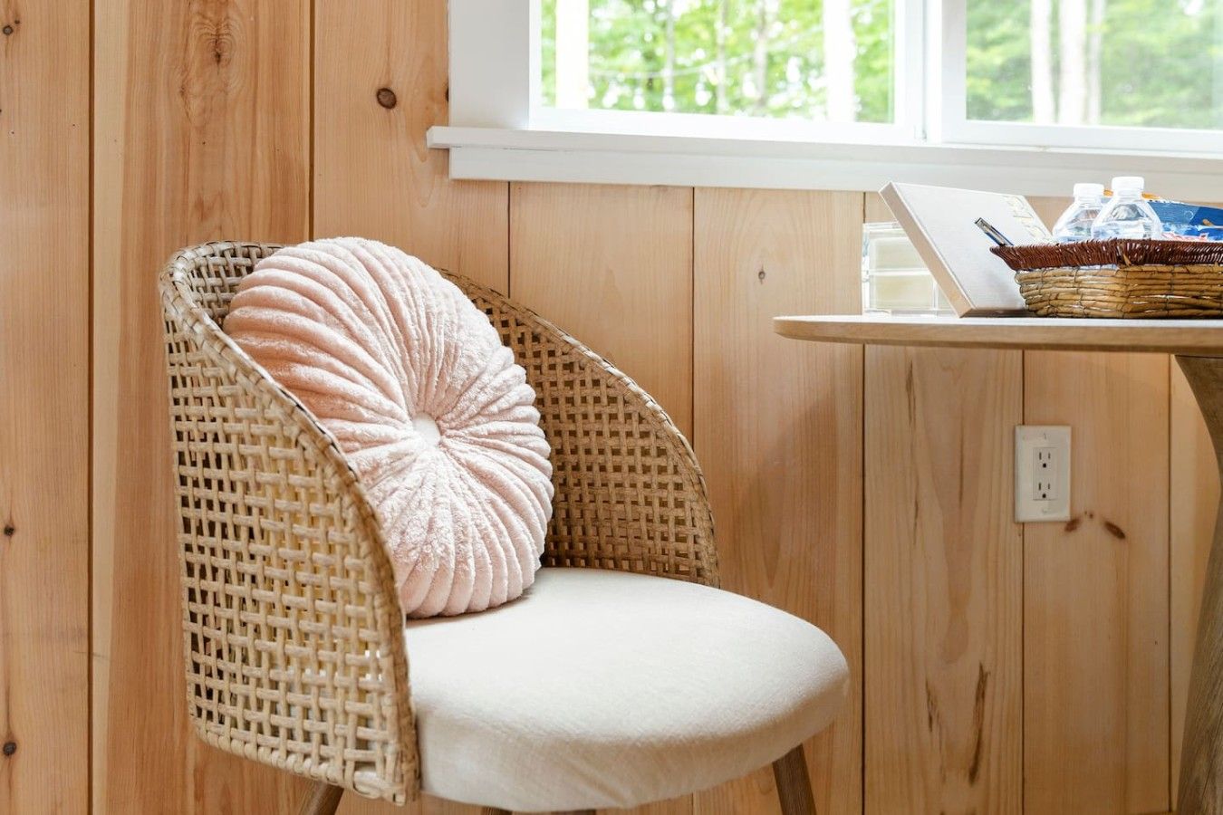 Una silla de mimbre con asiento acolchado y un cojín decorativo, ubicada junto a una pared de madera y una mesa con una cesta que contiene botellas.