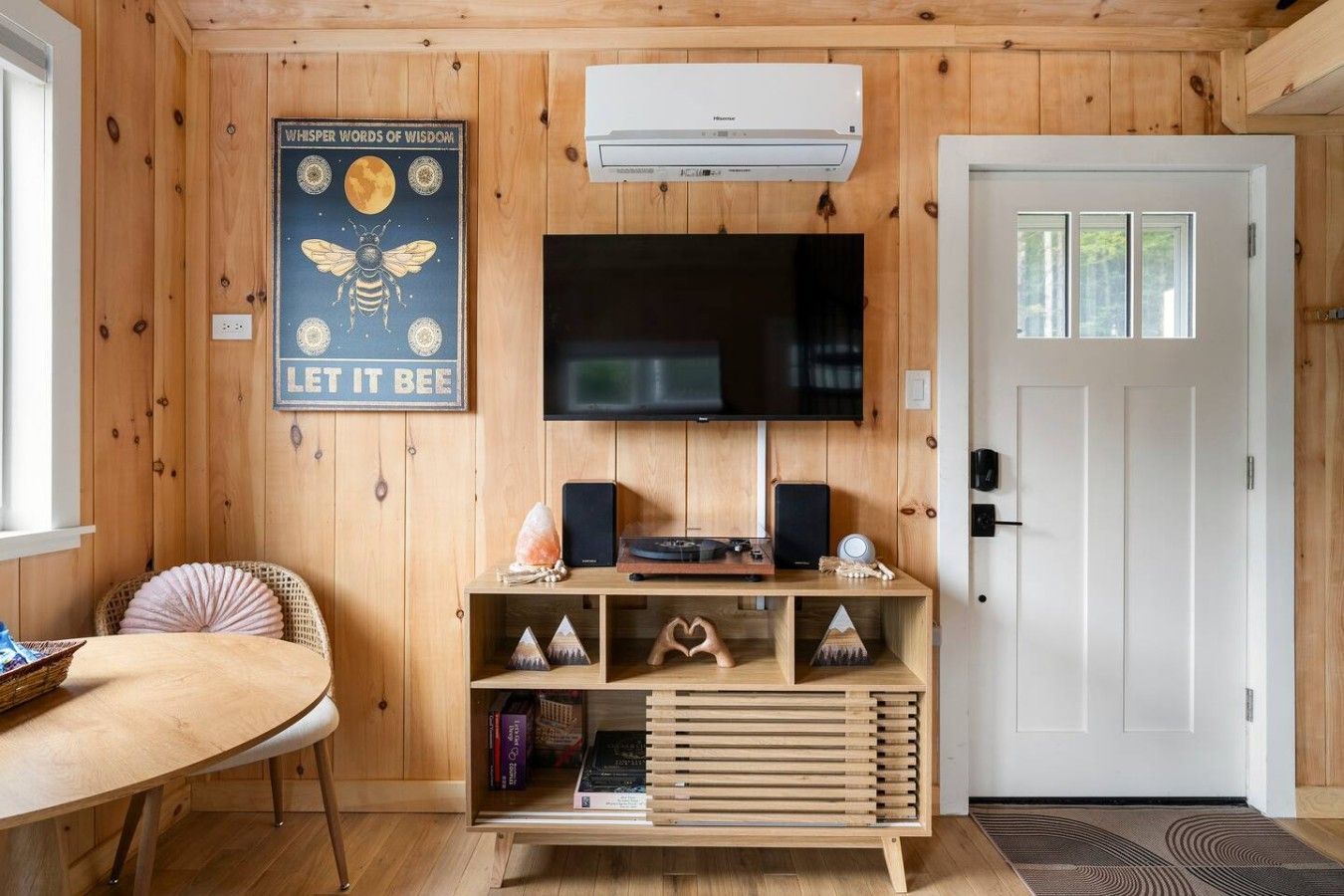 Una habitación con paredes de madera, un televisor de pantalla plana montado, un mueble de madera para entretenimiento con altavoces y adornos, una mesa redonda con una silla, y una puerta con ventanal.