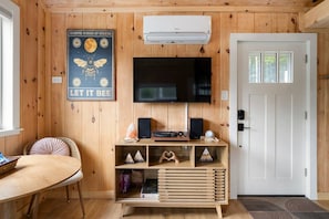 A room with wooden walls, a mounted flat-screen TV, a wooden entertainment unit with speakers and decorative items, a round table with a chair, and a door with a window.